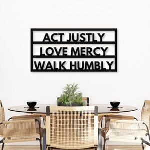 May include: Black metal wall art with the words "ACT JUSTLY, LOVE MERCY, WALK HUMBLY" in a modern font. The artwork is displayed above a dining table with woven chairs and a plant centerpiece, creating a minimalist aesthetic.