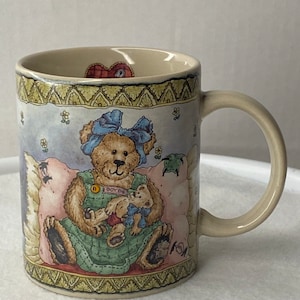 Puede incluir: Una taza de cerámica con un fondo azul claro que presenta un oso de peluche con un lazo azul y un vestido verde y blanco. El oso sostiene un oso de peluche más pequeño. La taza tiene un borde amarillo y verde con un diseño de corazón.