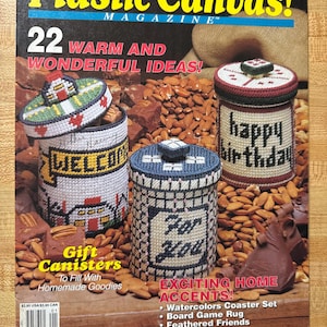 Könnte beinhalten: Das Cover des Magazins "Plastic Canvas!" zeigt drei handgefertigte Geschenkbehälter. Die zylindrischen Behälter sind mit Nadelspitzendesigns verziert. Einer sagt "Welcome", ein anderer "For You" und der dritte "Happy Birthday". Das Cover enthält auch Text über Wohnaccessoires.