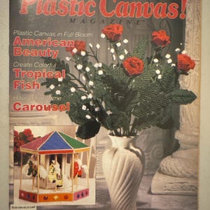 May include: A magazine cover featuring the title "Plastic Canvas!" in red, with a bouquet of red roses and greenery in a cream-colored vase. The cover also includes text about plastic canvas projects, including "American Beauty" and "Carousel."