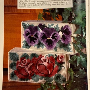 May include: Two doorstop cushions with floral embroidery. The top cushion features a purple pansy design, while the bottom cushion features a red rose design. Both cushions are white with green accents.