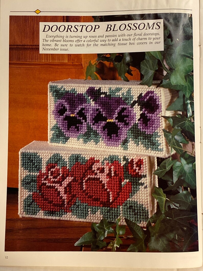 May include: Two doorstop cushions with floral embroidery. The top cushion features a purple pansy design, while the bottom cushion features a red rose design. Both cushions are white with green accents.