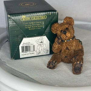 May include: A brown bear figurine with a black collar, sitting on a white surface. The bear is part of The Boyds Collection, a range of handcrafted figurines.