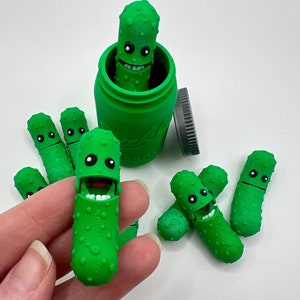May include: A green glass jar with a green lid filled with green pickle figurines. The jar has the word "Pickle" printed on the side. There are six pickle figurines in total, five of which are outside of the jar.