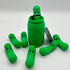 May include: A green glass jar with the word "Pickle" and "Mason" printed on it. The jar is filled with green pickle figurines with faces. There are also several green pickle figurines scattered around the jar.