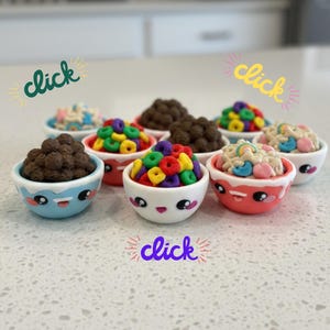 May include: A collection of small, decorative bowls filled with colorful cereal. The bowls are white with cute, cartoonish faces and are in various colors. The cereal includes chocolate puffs, colorful loops, and marshmallow shapes. The word "click" is written in different colors.