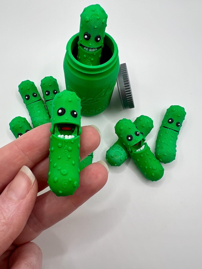 May include: A green glass jar with a green lid filled with green pickle figurines. The jar has the word "Pickle" printed on the side. There are six pickle figurines in total, five of which are outside of the jar.