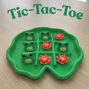 May include: A green Tic-Tac-Toe game set. The game board is shaped like a lily pad and features frog and flower game pieces. The text "Tic-Tac-Toe" is printed in green above the game board.