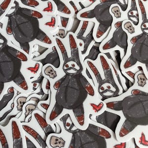 Puede incluir: Una colección de pegatinas con un conejo de dibujos animados con un cuerpo gris oscuro, detalles en rojo y una calavera. Las pegatinas también incluyen pequeñas formas de corazón rojo y calaveras adicionales. Las pegatinas son blancas con un acabado brillante.