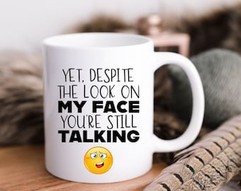 May include: White ceramic coffee mug with black text that reads "Yet, despite the look on my face you're still talking" and a yellow cartoon face with a confused expression.