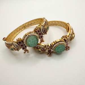 May include: A pair of gold bangles with intricate designs and green and red gemstones. The bangles are decorated with small white beads and have a carved green stone in the center.