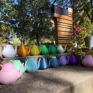 May include: A collection of colorful teapots in various shades, including pink, blue, green, yellow, and purple. The teapots are arranged on a stone surface, with a mailbox and greenery in the background. Each teapot has a handle and spout.