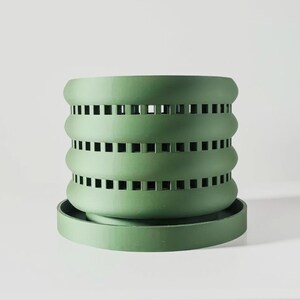 May include: A green plastic planter with a decorative, geometric design. The planter has a wide, flat base and a cylindrical body with multiple layers of square cutouts.