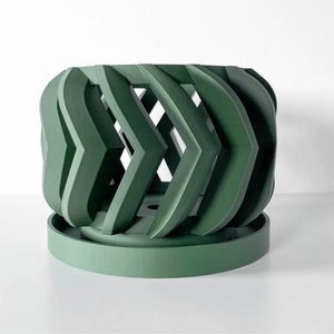 May include: A green ceramic planter with a geometric design. The planter has a cylindrical base and a decorative top with a series of interlocking, curved shapes.