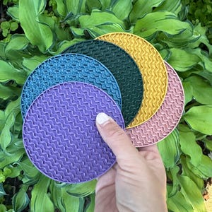 May include: Four round, textured silicone coasters in purple, teal, green, and yellow. The coasters are stacked on top of each other.