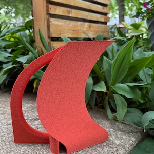 May include: A textured, coral-colored abstract sculpture with a curved design. The sculpture is placed outdoors on a stone surface, with greenery and a wooden structure in the background. The artwork has a unique, modern aesthetic.