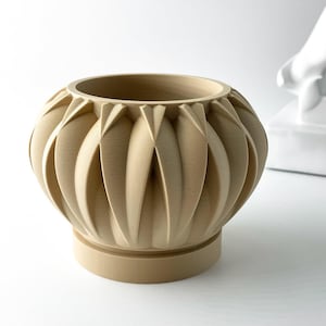May include: A beige ceramic planter with a unique geometric design. The planter has a wide, round base and a slightly flared top. The design features a series of overlapping, curved lines that create a textured, almost organic look.