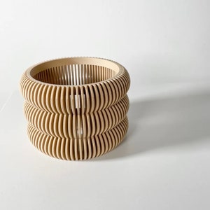 May include: A beige wooden planter with a unique, layered design. The planter is made of thin, curved slats that create a geometric pattern.