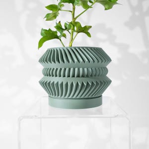May include: A sage green ceramic planter with a geometric design. The planter has a wide base and a narrow top, with a series of ridges and grooves that create a textured surface. The planter is sitting on a clear acrylic stand.