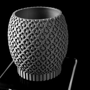 May include: A grayscale, cylindrical vase with a complex, repeating pattern across its surface. The vase sits on a clear, rectangular stand against a black background. The base of the vase has vertical lines, and the top is open.