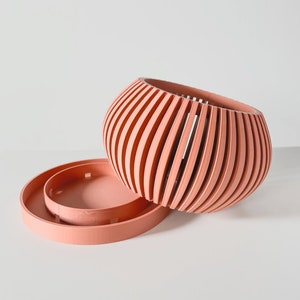May include: A set of two coral-colored ceramic planters. The larger planter has a round, ribbed design. The smaller planter is a flat, round disc.