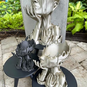 May include: Three silver and black decorative sculptures of a woman's head with a melted, dripping effect. The sculptures are displayed on black wooden stands.