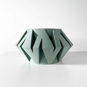 May include: A light green geometric ceramic planter with a unique folded design. The planter has a wide, round base and a narrow opening.
