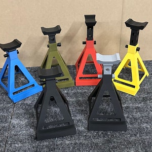May include: Six car jack stands in different colors: black, blue, gray, green, red, and yellow. The stands are made of metal and have a triangular base.