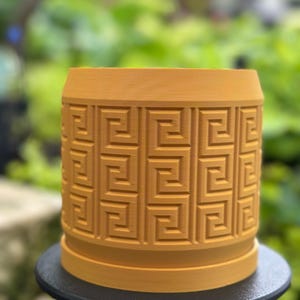 May include: A mustard yellow ceramic planter with a geometric pattern. The planter is sitting on a black tripod stand.