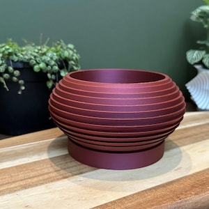 May include: A burgundy red, round, geometric planter with a ribbed design. The planter is made of a smooth, matte material and has a slightly raised base.