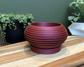 May include: A burgundy red, round, geometric planter with a ribbed design. The planter is made of a smooth, matte material and has a slightly raised base.