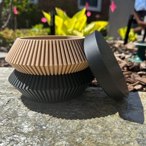 May include: A set of two nesting bowls, one black and one beige, with a ribbed design. The bowls are made of a smooth, hard material and have a rounded shape.