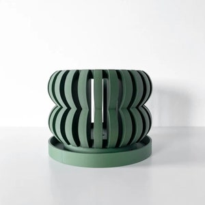 May include: A green planter with a unique, geometric design. The planter has a wide, round base and a tall, cylindrical body with a series of curved, overlapping panels.
