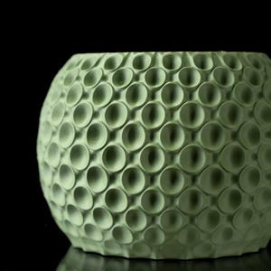 May include: A light green, spherical decorative bowl with a textured surface. The surface is covered in a pattern of evenly spaced, recessed circles. The bowl is set against a black background, highlighting its unique design.