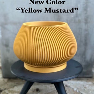 May include: A yellow mustard colored ceramic planter with a ribbed design sits on a black three-legged stand. The text "New Color "Yellow Mustard"" is displayed above the planter.