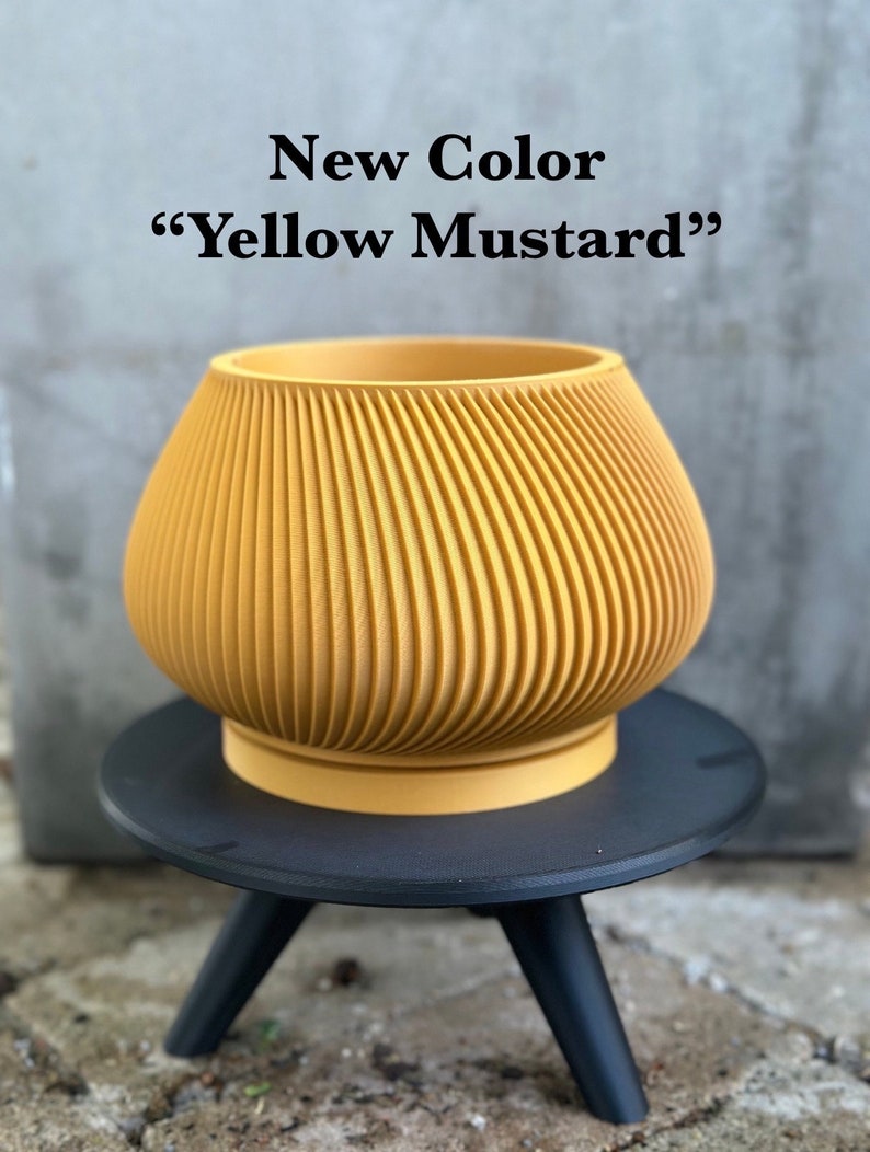 May include: A yellow mustard colored ceramic planter with a ribbed design sits on a black three-legged stand. The text "New Color "Yellow Mustard"" is displayed above the planter.