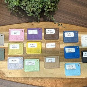 May include: A collection of 18 small square color swatches with different matte paint colors. The swatches are arranged in rows on a wooden surface. The colors include muted blue, muted white, muted purple, muted green, yellow marble, yellow, wood brown, denim, white, blue, ash gray, marble, mint green, brown, and light blue.