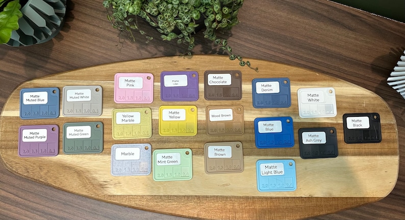 May include: A collection of 18 small square color swatches with different matte paint colors. The swatches are arranged in rows on a wooden surface. The colors include muted blue, muted white, muted purple, muted green, yellow marble, yellow, wood brown, denim, white, blue, ash gray, marble, mint green, brown, and light blue.