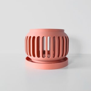 May include: A coral pink ceramic planter with a decorative, open-work design. The planter has a matching saucer.