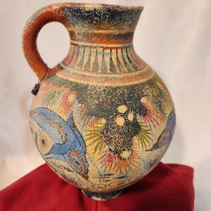 May include: A hand-painted ceramic jug with a rounded body and a handle. The jug features a dark blue background with colorful illustrations of birds and fish. The handle is red, and the rim has a decorative pattern.
