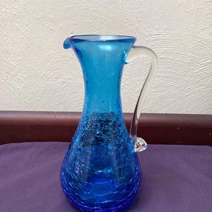 May include: A vibrant blue glass pitcher with a crackle finish. The pitcher has a curved handle and a spout for easy pouring. The glass is a deep, rich blue color, perfect for serving beverages.