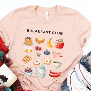 May include: A pink t-shirt with the text "Breakfast Club" printed on the front. The shirt features a graphic design of various breakfast foods, including waffles, eggs, bacon, donuts, and coffee.