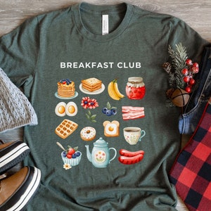 May include: A green t-shirt with the text "Breakfast Club" printed on the front. The shirt features a variety of breakfast foods, including waffles, eggs, bacon, and coffee.
