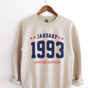 May include: A beige sweatshirt with the text "JANUARY 1993 - LIMITED EDITION" in navy blue and red. The design includes red stars above the word "JANUARY". The sweatshirt is hanging on a wooden hanger.