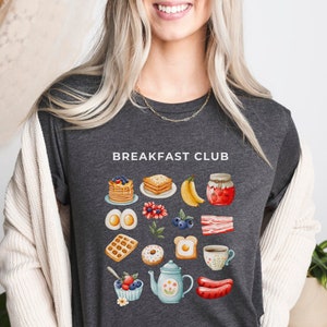 May include: A dark gray t-shirt with the text "Breakfast Club" printed on the front. The shirt features a colorful illustration of breakfast foods, including waffles, eggs, bacon, pancakes, and coffee.