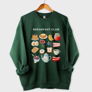 May include: A dark green sweatshirt with the text "Breakfast Club" printed on the front. The design features a variety of breakfast foods, including waffles, eggs, bacon, and fruit.