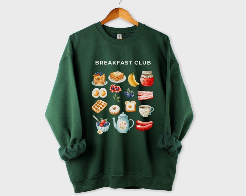 May include: A dark green sweatshirt with the text "Breakfast Club" printed on the front. The design features a variety of breakfast foods, including waffles, eggs, bacon, and fruit.