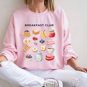 May include: A pink sweatshirt with the text "Breakfast Club" printed on the front. The design features a variety of breakfast foods, including pancakes, waffles, eggs, bacon, and fruit.