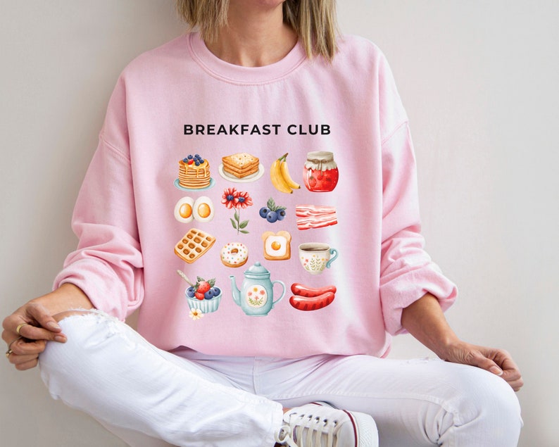 May include: A pink sweatshirt with the text "Breakfast Club" printed on the front. The design features a variety of breakfast foods, including pancakes, waffles, eggs, bacon, and fruit.