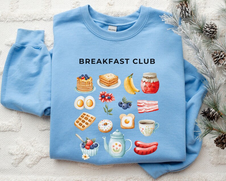 May include: Light blue sweatshirt with the text "BREAKFAST CLUB" in black. The sweatshirt features a variety of breakfast foods, including pancakes, waffles, bacon, eggs, and a donut, all illustrated in a watercolour style.